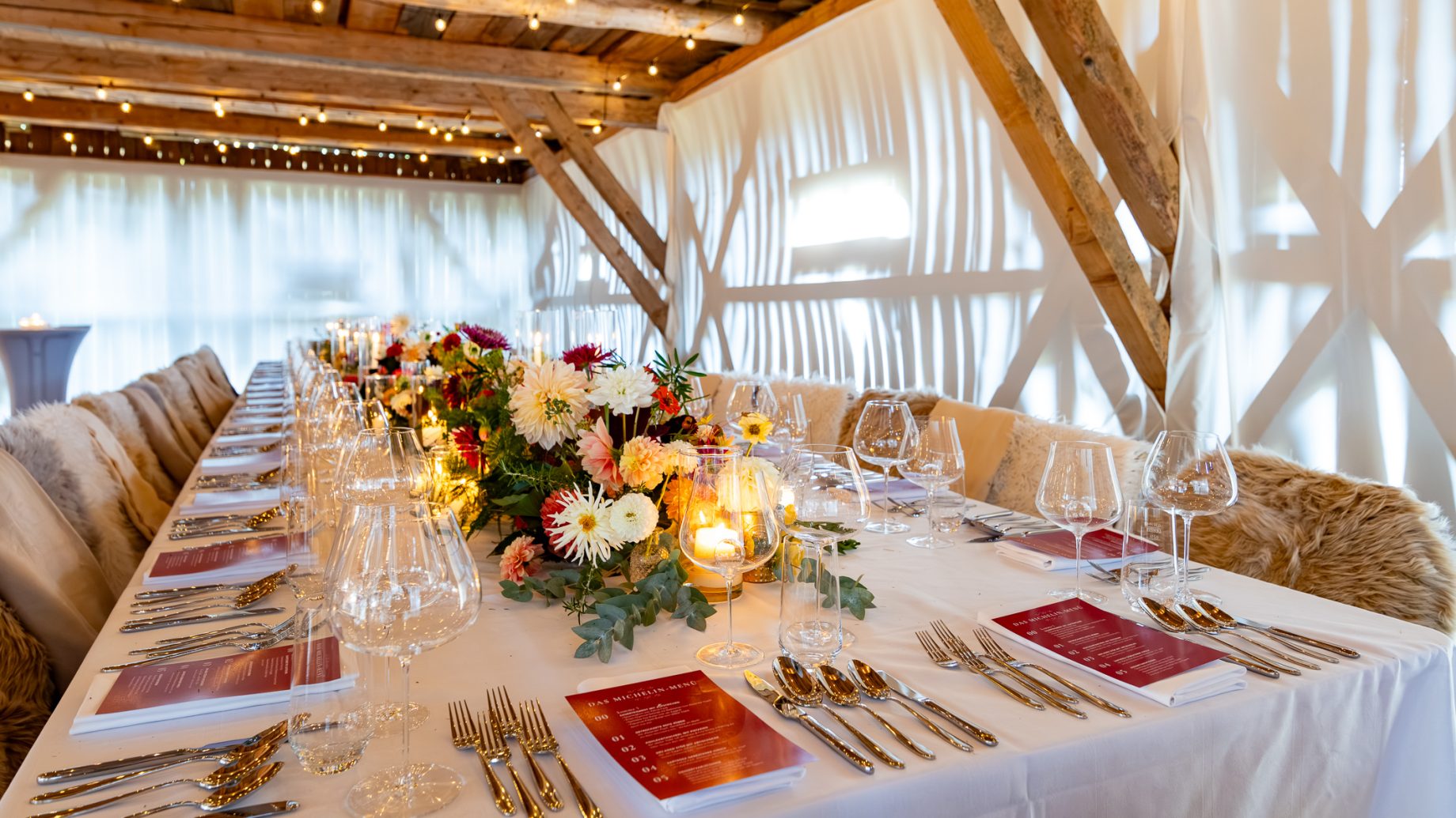 Ein langer Banketttisch, gedeckt für „Der gedeckte Ort“, Band 3, mit Blumengestecken, Kerzen, Weingläsern, Besteck und roten Menükarten.