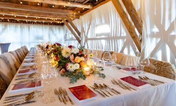 Ein langer Banketttisch, gedeckt für „Der gedeckte Ort“, Band 3, mit Blumengestecken, Kerzen, Weingläsern, Besteck und roten Menükarten.