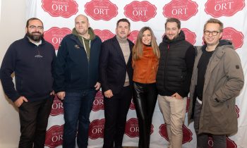 Sechs Personen stehen in einer Reihe vor einem mit dem Logo versehenen Hintergrund für „Der gedeckte Ort, Band 3“.