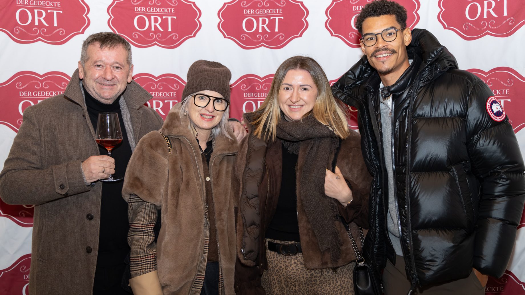 Vier Personen posieren vor einer Kulisse mit dem Schriftzug „Der gedeckte Ort, Band 3“ für ein Foto.