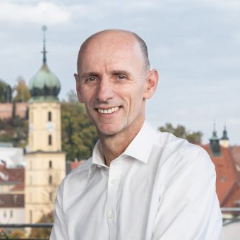 Ein lächelnder Mann mittleren Alters in einem weißen Hemd mit Kragen steht vor einem Kirchturm und den Dächern der Stadt.