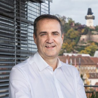 Ein Mann mittleren Alters in einem weißen Hemd steht vor einer Stadtkulisse.