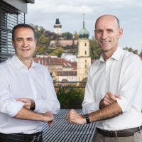 Zwei Männer in weißen Hemden stehen auf einem Balkon mit Blick auf die Stadt Graz in Österreich.