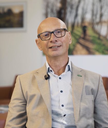 Christian Contola, ein kahlköpfiger Mann mit Brille, der einen beigen Blazer und ein gemustertes Hemd trägt, lächelt in die Kamera.