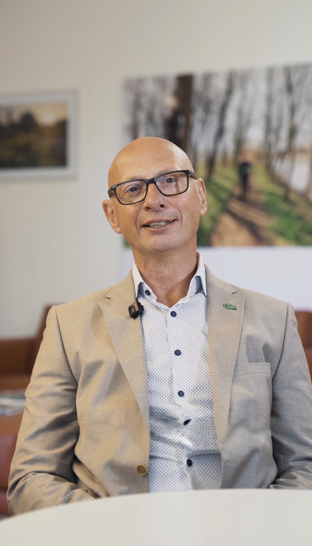 Christian Contola, ein kahlköpfiger Mann mit Brille, der einen beigen Blazer und ein gemustertes Hemd trägt, lächelt in die Kamera.