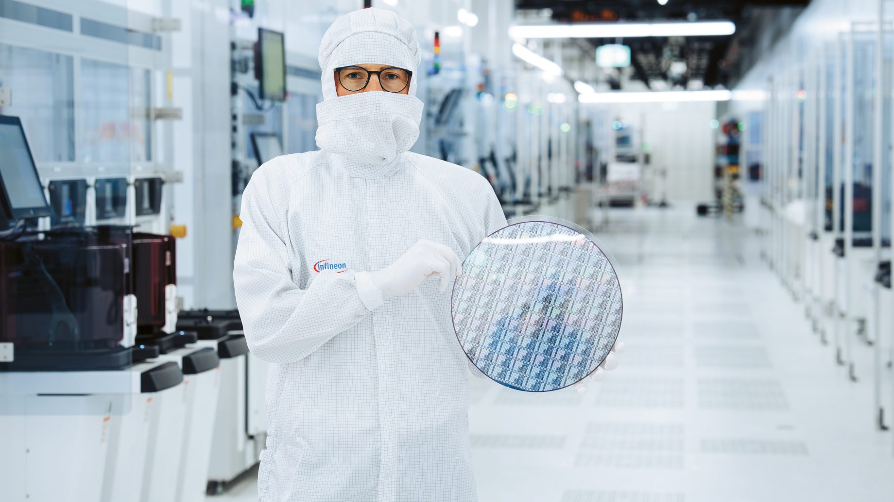 Ein Techniker in einem weißen Reinraumanzug hält in einer Halbleiterfertigungsanlage einen Siliziumwafer in der Hand.