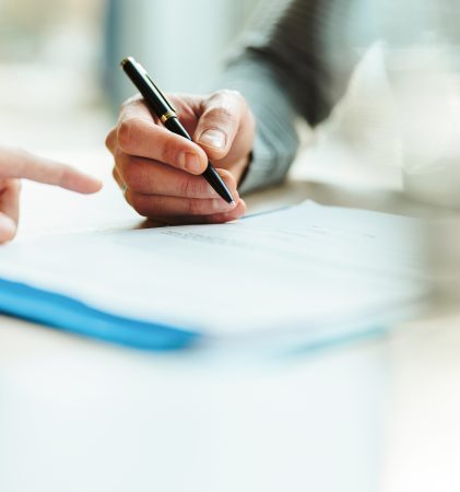Eine Person zeigt auf ein Dokument, während eine andere Person einen Stift bereit hält, um es zu unterschreiben.