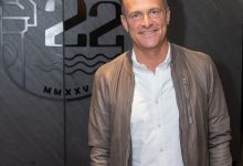 Matthias Malmedie steht in einem weißen Hemd und einer hellbraunen Jacke vor einer Wand von J22 Graz, auf der ein Logo mit der Zahl 22 und den römischen Ziffern MMXXV zu sehen ist.