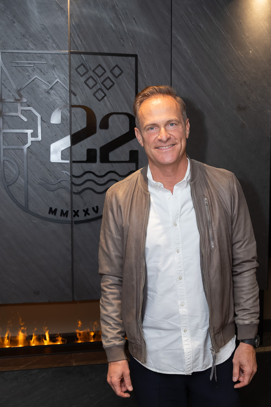 Matthias Malmedie steht in einem weißen Hemd und einer hellbraunen Jacke vor einer Wand von J22 Graz, auf der ein Logo mit der Zahl 22 und den römischen Ziffern MMXXV zu sehen ist.
