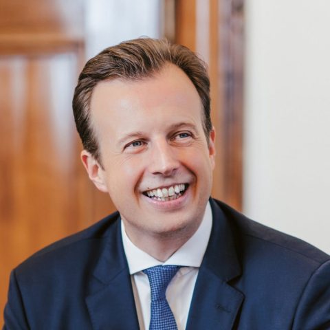 Ein lächelnder Mann in einem dunkelblauen Anzug und einer blauen Krawatte sitzt vor einer Holzwand und blickt leicht nach rechts.