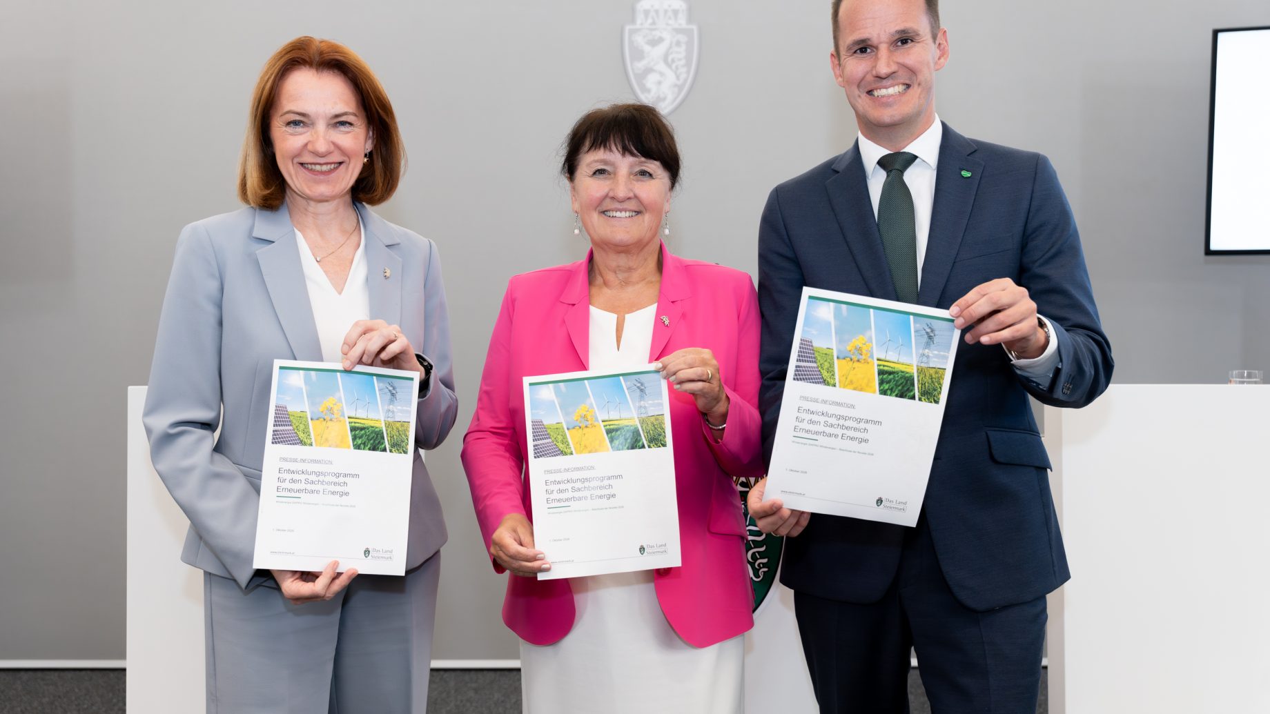 Drei lächelnde Personen in Geschäftskleidung halten Dokumente in den Händen und feiern vor einer grauen Wand die Erweiterung des Energieportfolios in der Steiermark: Windkraft und Solarstrom.