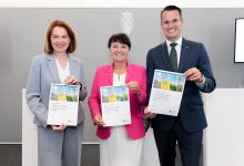 Drei lächelnde Personen in Geschäftskleidung halten Dokumente in den Händen und feiern vor einer grauen Wand die Erweiterung des Energieportfolios in der Steiermark: Windkraft und Solarstrom.