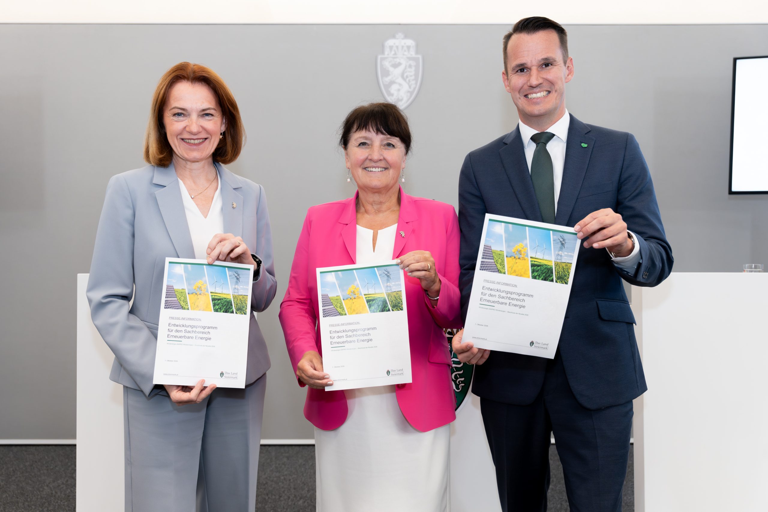 Drei lächelnde Personen in Geschäftskleidung halten Dokumente in den Händen und feiern vor einer grauen Wand die Erweiterung des Energieportfolios in der Steiermark: Windkraft und Solarstrom.