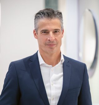 Ein Mann mittleren Alters mit graumeliertem Haar, der einen marineblauen Blazer und ein weißes Hemd trägt.