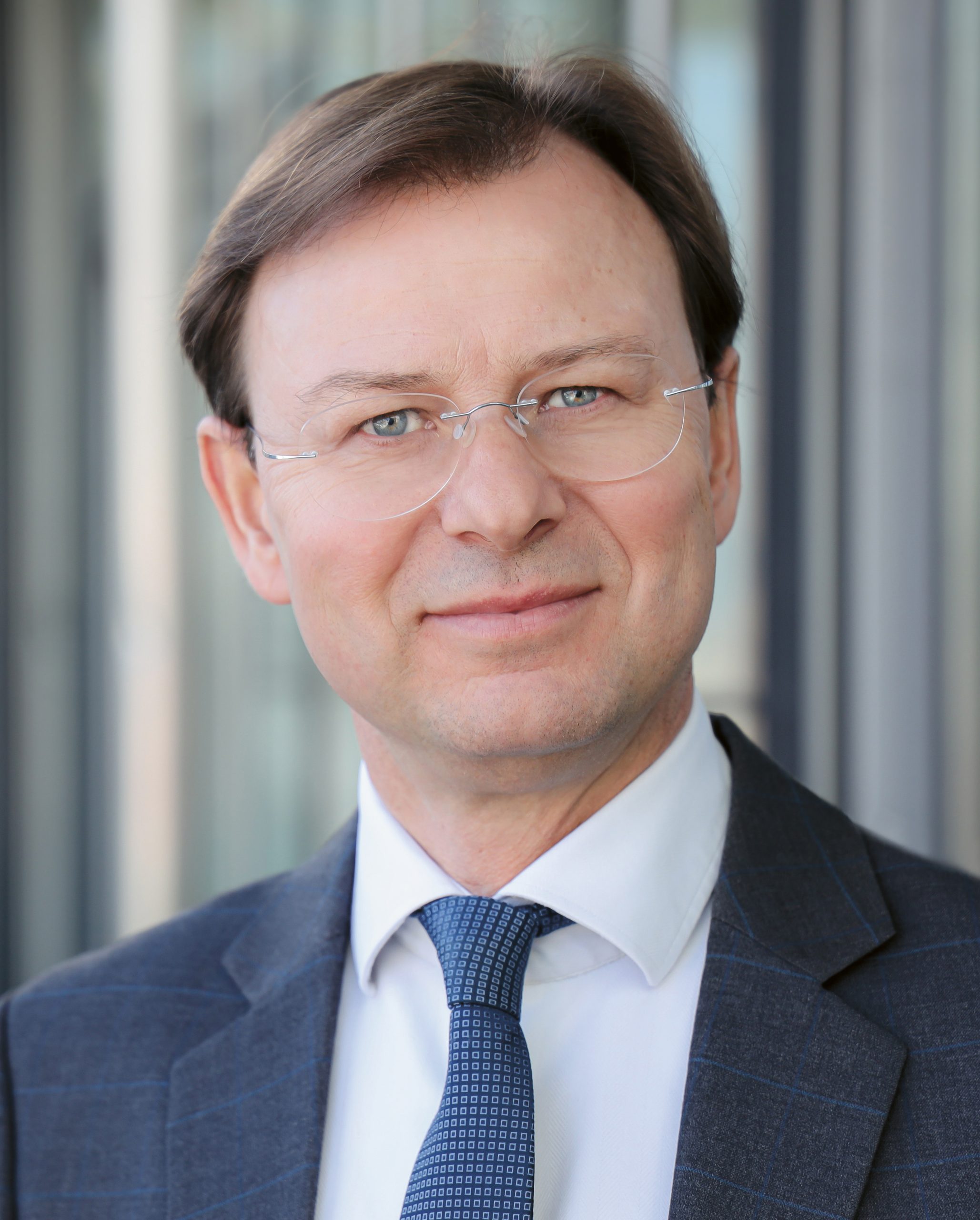 Ein Mann mittleren Alters in Anzug, weißem Hemd und blau gemusterter Krawatte, der eine randlose Brille trägt.