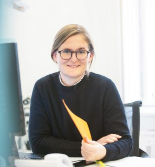 Eine lächelnde Frau mit Brille, die einen schwarzen Pullover trägt, sitzt an einem Schreibtisch und hält eine orangefarbene Mappe in der Hand.