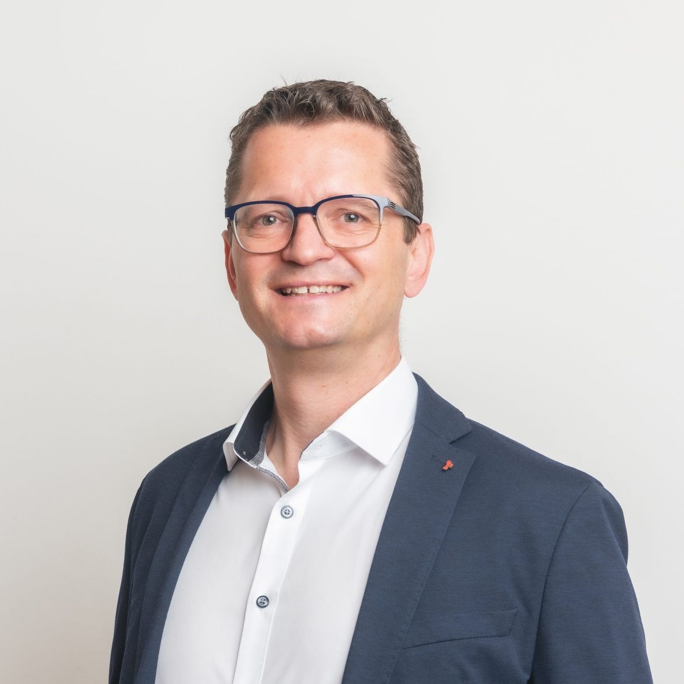 Ein lächelnder Mann in einem dunklen Blazer, einem weißen Hemd und einer Brille steht vor einem schlichten weißen Hintergrund.