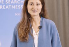 Eine Frau spricht vor einem blauen BDO-Banner mit der Aufschrift „WE SEARCH FOR GREATNESS“.