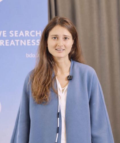 Eine Frau spricht vor einem blauen BDO-Banner mit der Aufschrift „WE SEARCH FOR GREATNESS“.