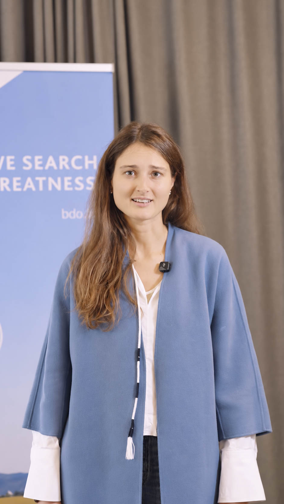 Eine Frau spricht vor einem blauen BDO-Banner mit der Aufschrift „WE SEARCH FOR GREATNESS“.