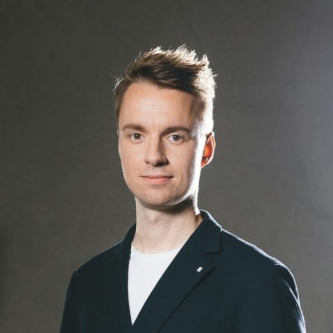 Ein Mann in einem Blazer und einer hellen Hose steht in einem Studio, hinter ihm leuchten helle Lampen und Scheinwerfer, und er posiert selbstbewusst mit einer Hand in der Hosentasche.