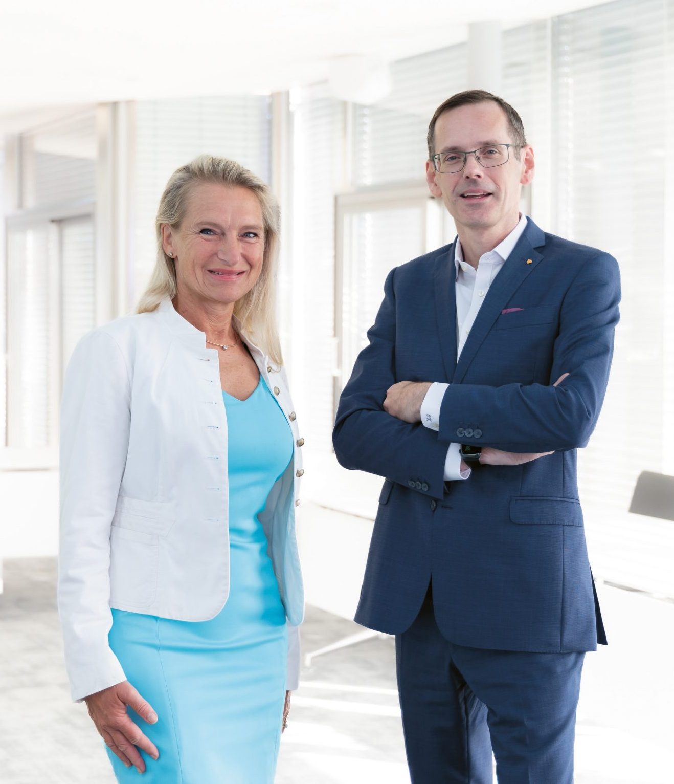 Eine blonde Frau in einer weißen Jacke und einem blauen Kleid steht neben einem Mann in einem blauen Anzug, der die Arme verschränkt hat.