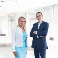 In einem hellen, modernen Büro steht eine berufstätige Frau in einem hellblauen Kleid und einer weißen Jacke neben einem Mann in einem marineblauen Anzug.