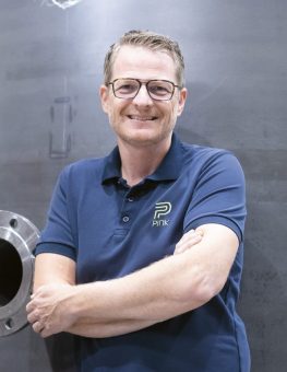 Vor einem grauen Industriehintergrund steht eine lächelnde Person mit Brille und kurzen Haaren, die ein marineblaues Poloshirt mit dem PINK-Logo trägt.