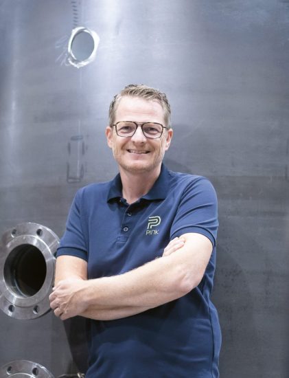 Eine Person mit Brille und einem blauen Polohemd steht lächelnd mit verschränkten Armen vor einer großen Industrieanlage in einer Fabrikumgebung.