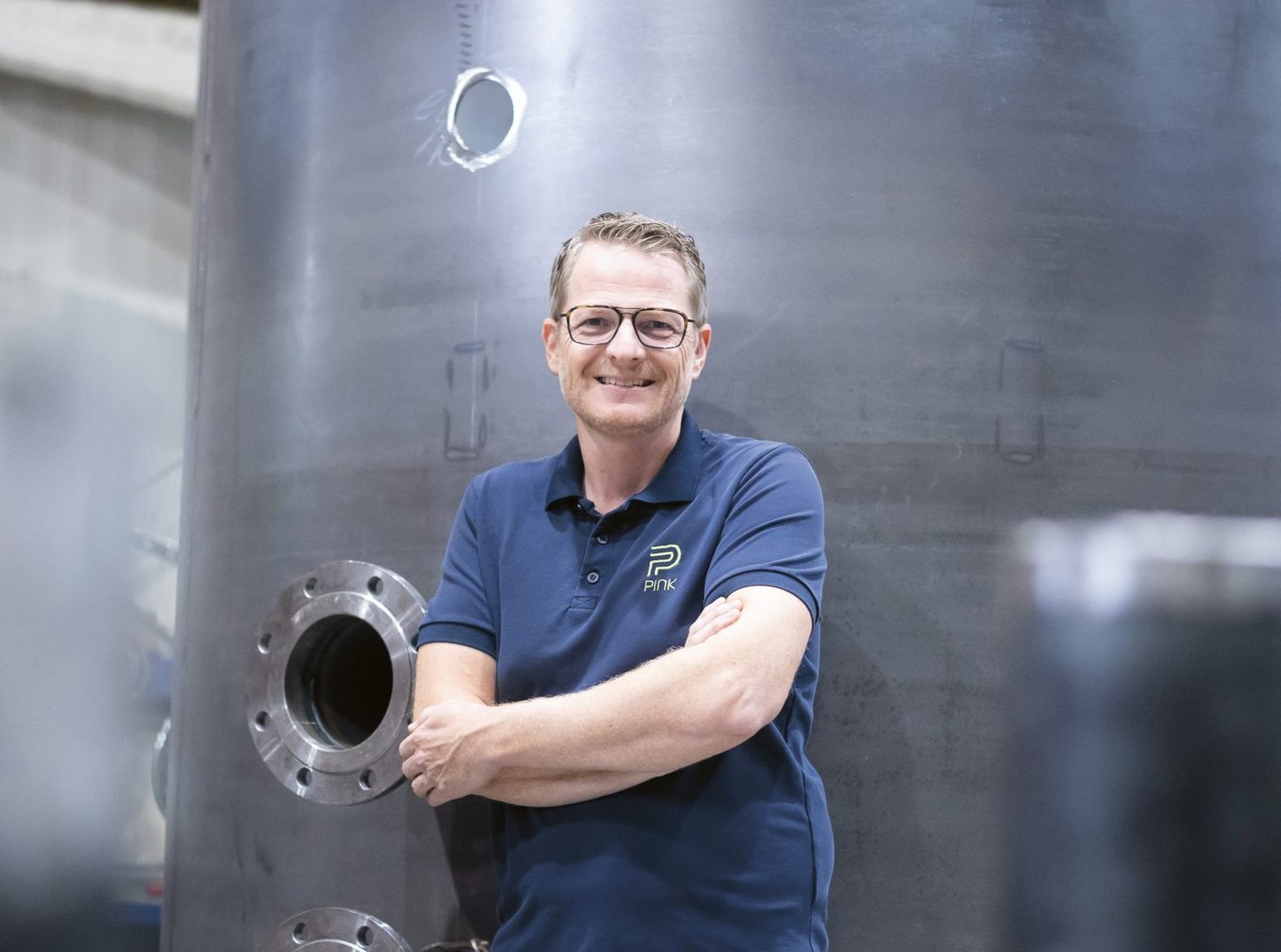 Eine Person mit Brille und einem blauen Polohemd steht lächelnd mit verschränkten Armen vor einer großen Industrieanlage in einer Fabrikumgebung.