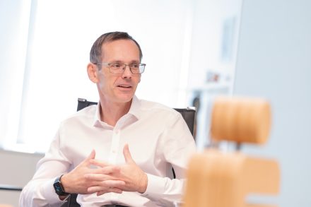 Ein Mann mittleren Alters mit Brille, der ein weißes Hemd trägt, sitzt mit gefalteten Händen an einem Schreibtisch.
