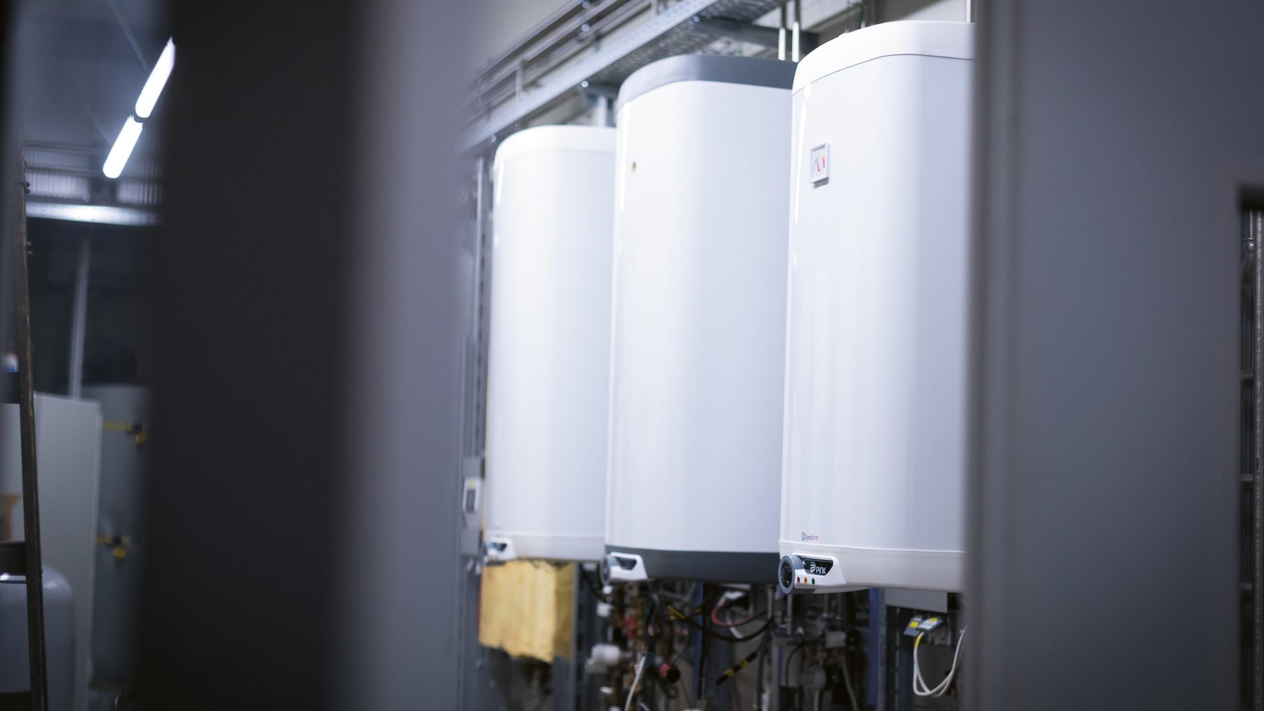 In einem Industrie- oder Hauswirtschaftsraum sind drei große weiße Warmwasserbereiter oder Heizkessel in einer Reihe aufgestellt, wobei die Rohrleitungen und Kabel sichtbar sind.