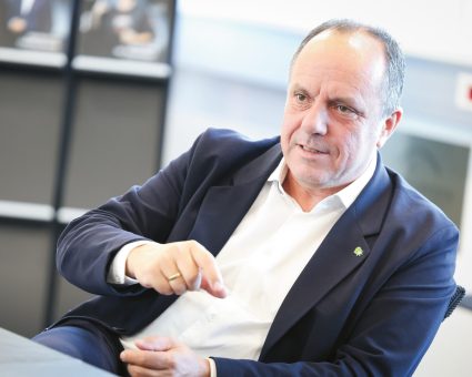 Ein Mann mittleren Alters in einem marineblauen Blazer und einem weißen Hemd spricht während einer Besprechung und untermalt seine Worte mit Gesten.