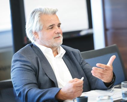 Ein Mann mittleren Alters mit grauen Haaren, der einen Blazer trägt, sitzt an einem Tisch und gestikuliert, während er bei einer Besprechung spricht.