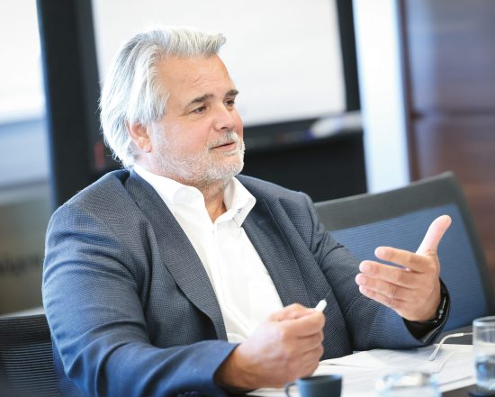 Ein Mann mittleren Alters mit grauen Haaren, der einen Blazer trägt, sitzt an einem Tisch und gestikuliert, während er bei einer Besprechung spricht.