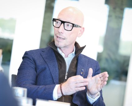 Ein kahlköpfiger Mann mit Brille in einem blauen Blazer spricht während einer Besprechung und gestikuliert dabei mit den Händen.