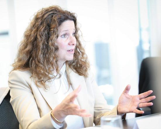 Eine Frau mit langen, welligen Haaren sitzt an einem Tisch und gestikuliert, während sie bei einer Besprechung spricht.