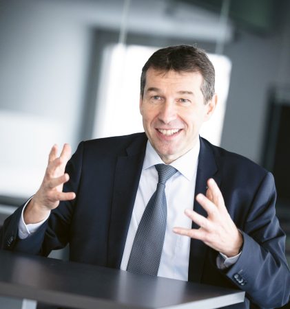 Ein lächelnder Mann in Anzug und Krawatte, der an einem Tisch sitzt und mit den Händen gestikuliert.