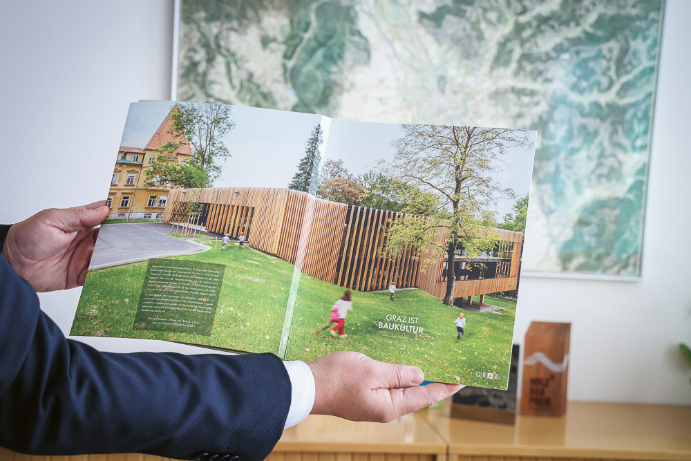 Eine Person, die ein aufgeschlagenes Buch in der Hand hält, auf dessen Seite ein modernes Holzgebäude in einer Parklandschaft zu sehen ist, zusammen mit dem Text „Graz ist Baukultur“.