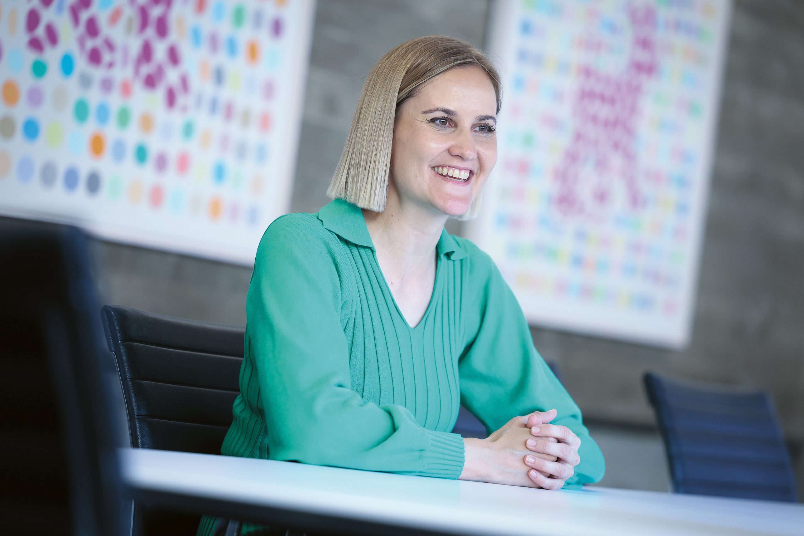 Eine lächelnde Frau mit schulterlangen blonden Haaren, die einen grünen Pullover trägt, sitzt an einem Tisch in einem Büro.