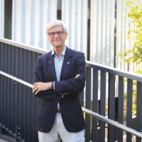 Ein Mann mit weißem Haar und Brille, der einen marineblauen Blazer und eine helle Hose trägt, steht mit verschränkten Armen auf einem Gehweg.