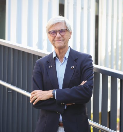 Ein Mann mit weißem Haar und Brille, der einen marineblauen Blazer und eine helle Hose trägt, steht mit verschränkten Armen auf einem Gehweg.