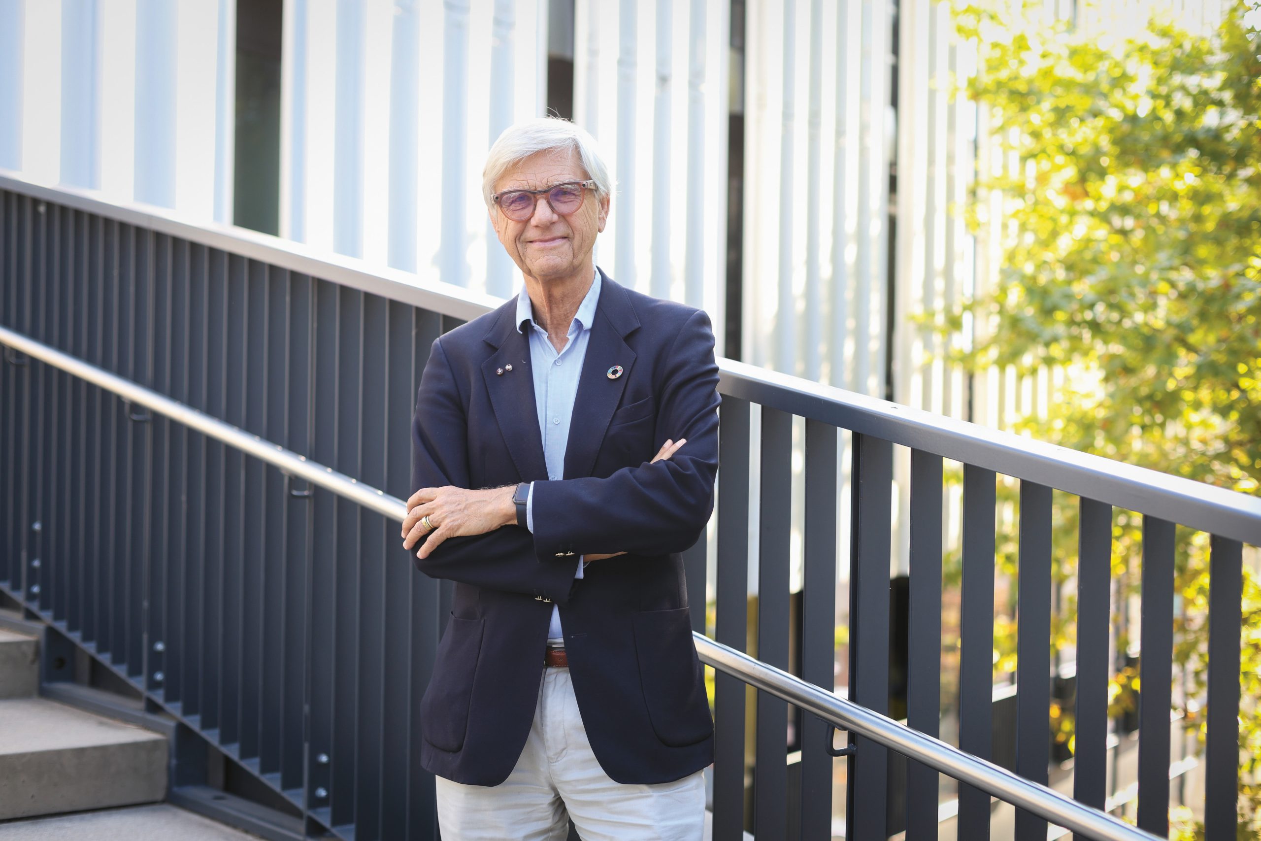 Ein Mann mit weißem Haar und Brille, der einen marineblauen Blazer und eine helle Hose trägt, steht mit verschränkten Armen auf einem Gehweg.