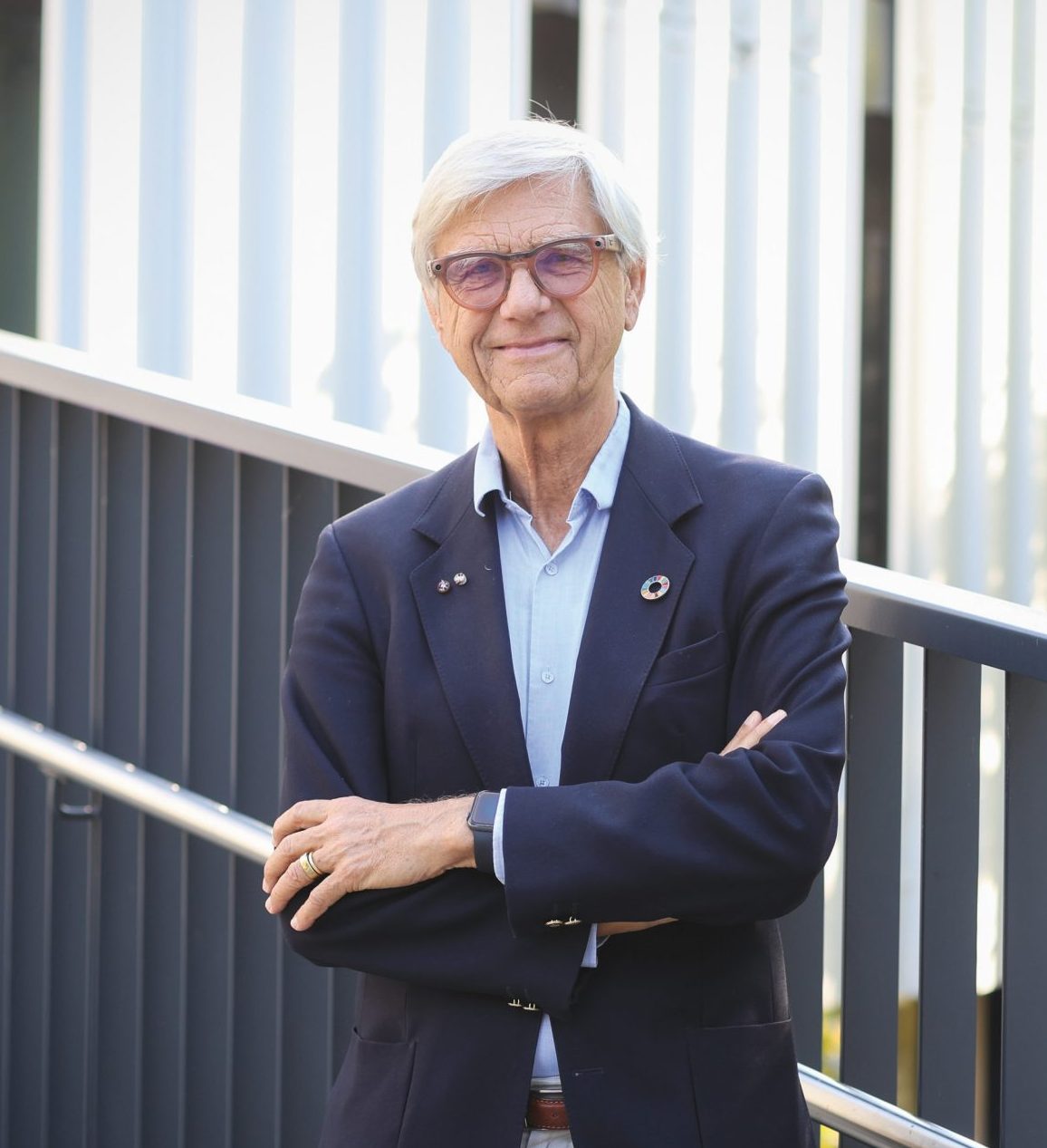 Ein Mann mit grauen Haaren und Brille, der einen marineblauen Blazer und ein hellblaues Hemd mit Knöpfen trägt, steht mit verschränkten Armen da.