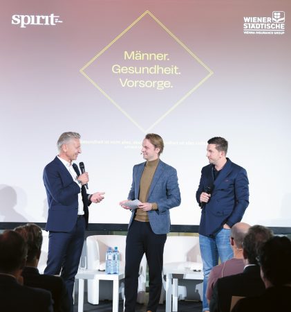 Drei Männer in Anzügen halten auf der Bühne einer Gesundheitsveranstaltung eine Rede, hinter ihnen ist auf einer Leinwand der Schriftzug „Männer. Gesundheit. Vorsorge.“ zu sehen.