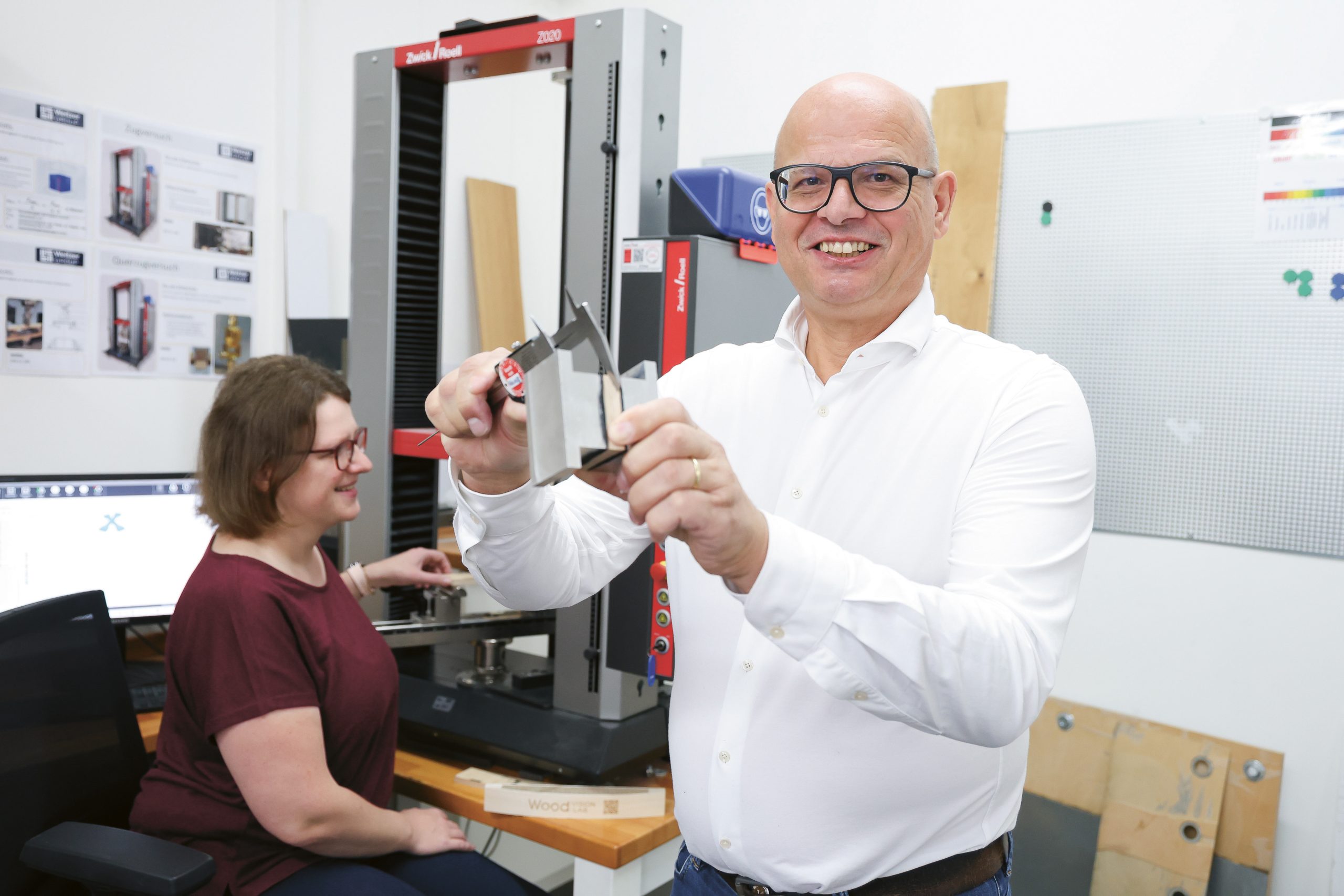 Ein Mann, der lächelnd ein Metallprüfstück mit einem Messschieber in der Hand hält, während im Hintergrund ein Kollege am Computer arbeitet und eine Materialprüfmaschine zu sehen ist.