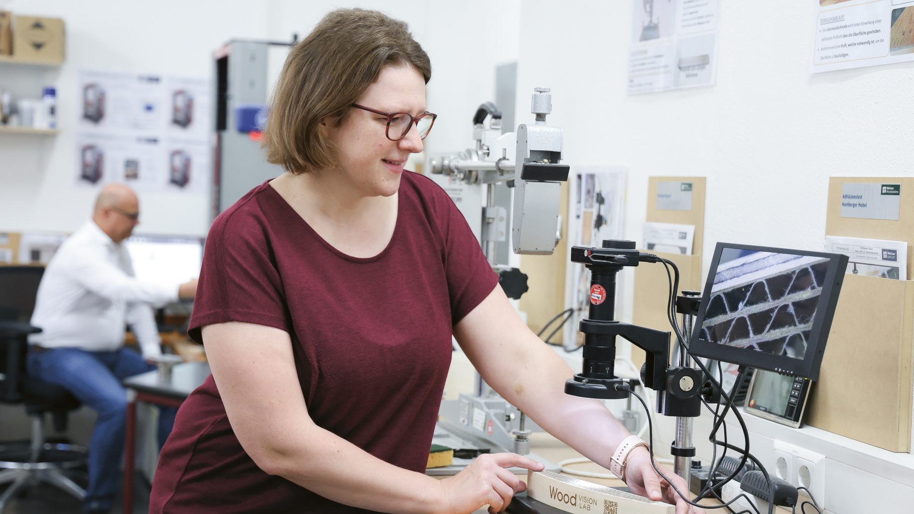 Eine Frau in einem burgunderroten Hemd untersucht in einem Labor mit einem Digitalmikroskop einen Holzblock, auf dem „Wood Vision Lab“ steht.