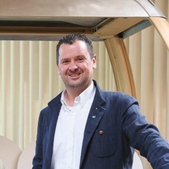 Ein lächelnder Mann, der ein weißes Hemd mit Kragen und einen dunkelblauen Blazer trägt, steht in einem Raum.