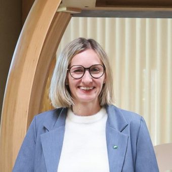 Eine Frau mit blondem Bob und Brille, die einen blauen Blazer über einem weißen Pullover trägt.