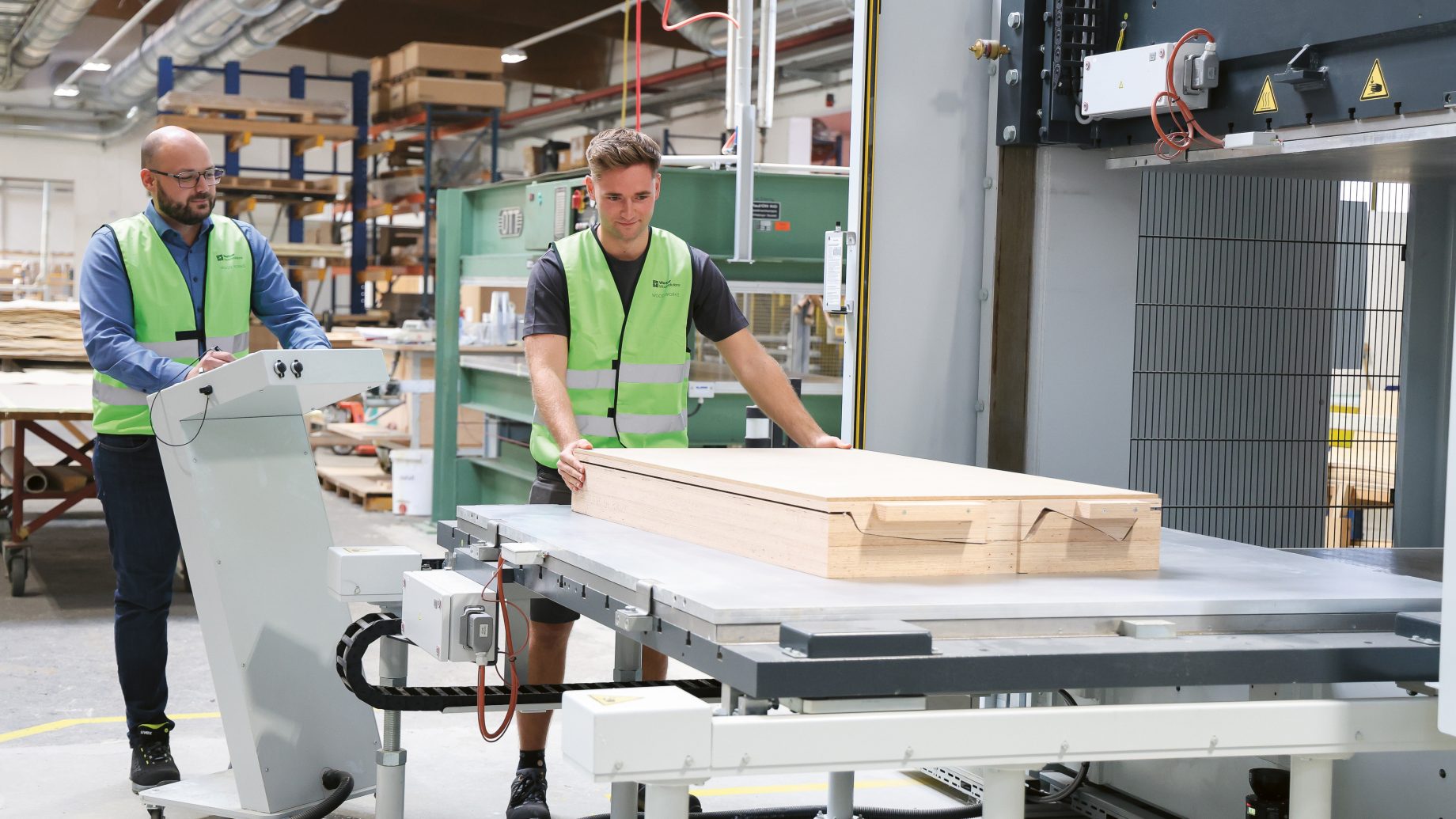 Zwei Arbeiter in Sicherheitswesten bedienen in einer Fabrik eine große industrielle Holzpresse.
